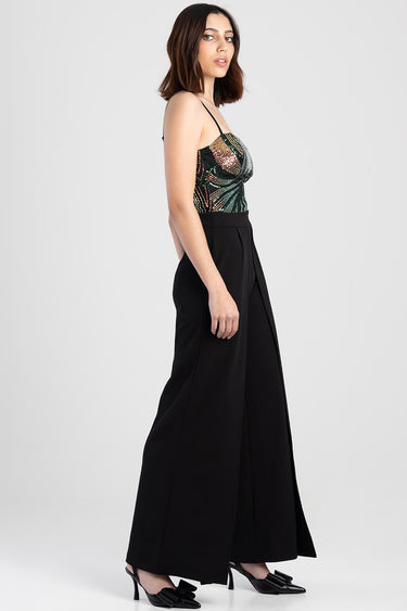 model wearing a green and gold sequined corset top with thin straps and black wide leg pants paired with black heeled mules