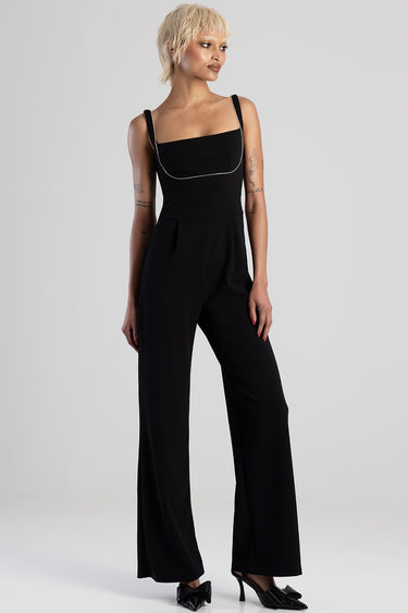 woman in black wideleg jumpsuit with square neckline and silver chain trim, standing with hands at sides, short blonde hair