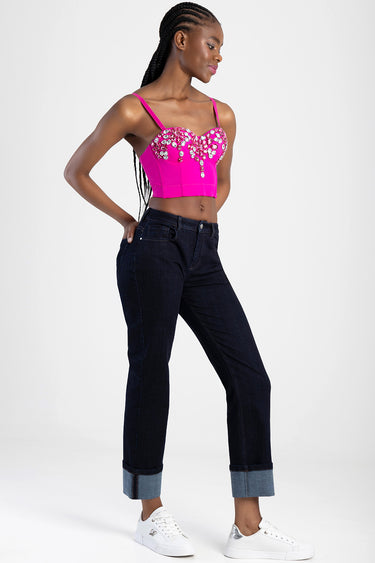 woman wearing a bright pink embellished bustier crop top with rhinestones and dark denim jeans rolled at the cuffs, white