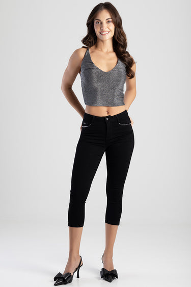 woman wearing a glittery grey sleeveless crop top and black capri jeans, standing with hands in pockets and smiling.