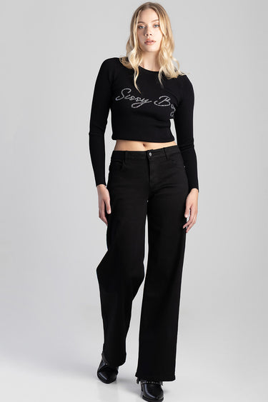 woman wearing black wide leg jeans and a cropped black knit top with silver script against a light grey backdrop