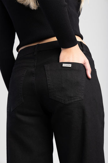Blue Jean Traders black wide leg jeans, back view with hand in back pocket and small metal logo tag on the right pocket