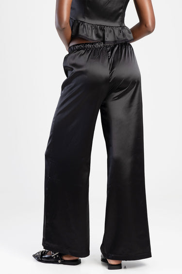back view of a model wearing black satin wide leg trousers with a gathered elastic waist