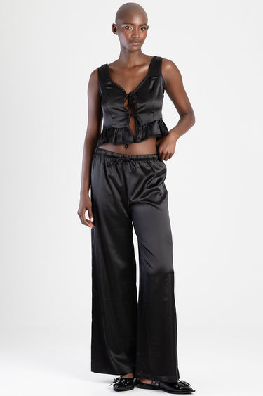 model in black satin crop top with tie front and ruffle hem, paired with wide leg satin trousers and black flats