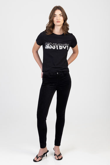 woman wearing black skinny pants and a black t shirt with a silver sequin logo, standing with hands on hips in sandals