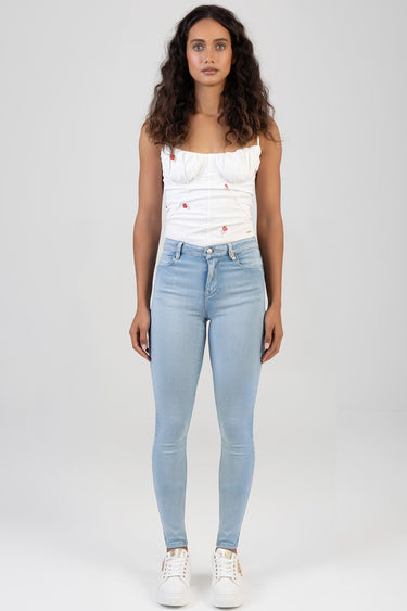 woman wearing light blue skinny jeans by blue jean traders and a white embroidered top, standing against a neutral backdrop