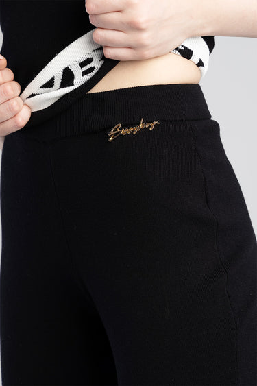 close up of black knit pants with a gold script logo at the hip and a ribbed waistband, as a black and white top is pulled up