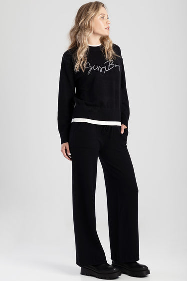 woman wearing a black knit sweater with white hem trim and cursive logo, paired with black wide‑leg pants and chunky boots
