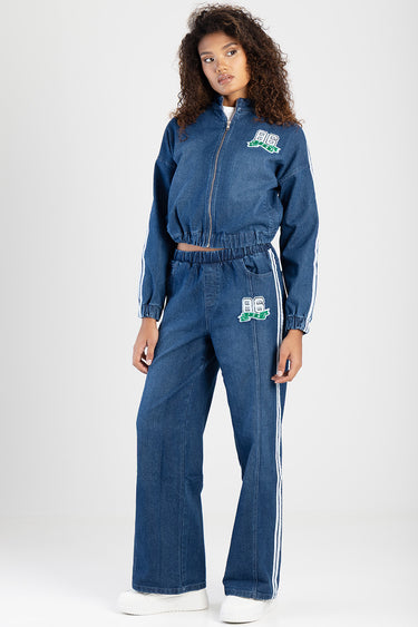 woman wearing a blue denim track suit featuring a zip jacket and wide leg trousers with white side stripes and embroidered