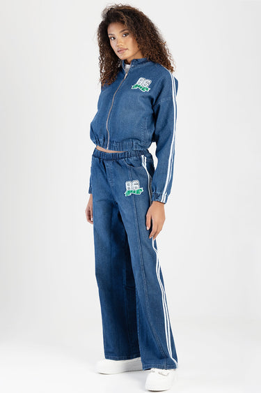 woman in a blue denim track suit with white side stripes, patch displaying eighty six on chest and hip, white sneakers