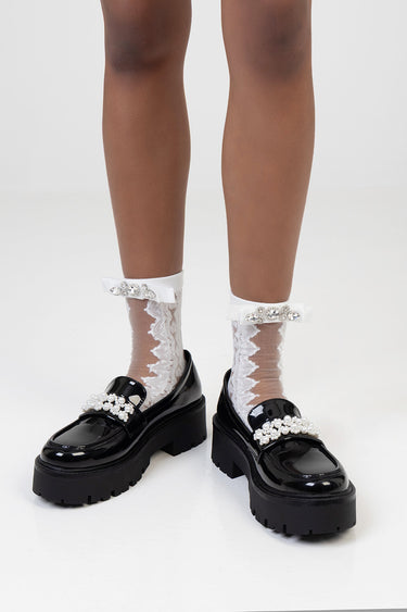 two legs wearing white lace ankle socks with bow and rhinestone ankle bands, and shiny black loafers with pearl