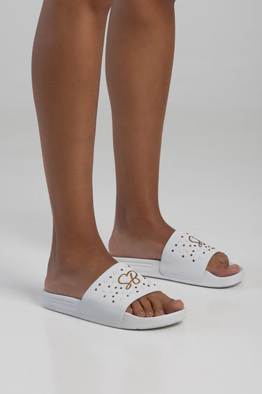 white slide sandals with a perforated upper and a small gold bow accent on the strap, worn on feet