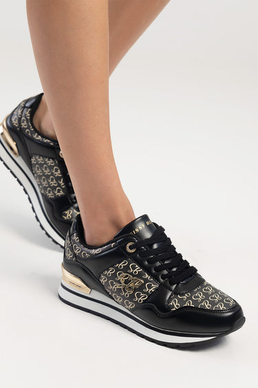 black sneakers with beige repeating motif and gold emblem, lace up, white and black sole on a model's foot