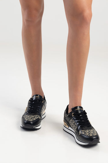 black sneakers with beige logo pattern, black laces, white midsole, and gold heel detail on a model's legs
