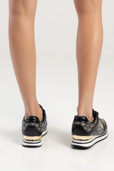 feet in black sneakers with a gold script pattern, white and black striped sole and gold heel trim