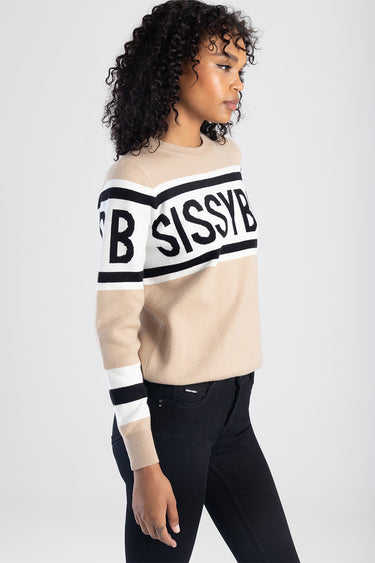 woman in beige knit top with bold black and white stripe across the chest and sleeves, standing in profile with black jeans