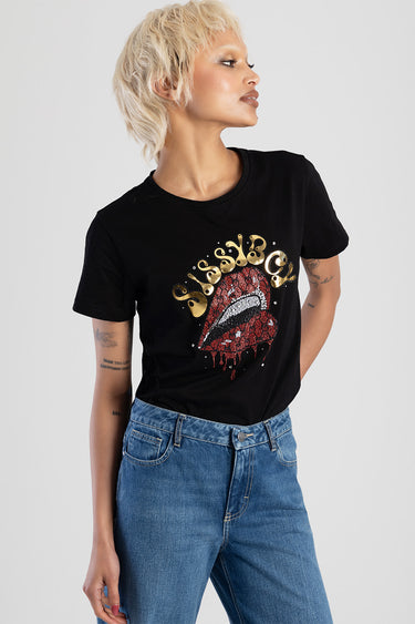 woman in black short sleeve top with a red sequined lips graphic and gold lettering, paired with blue denim jeans