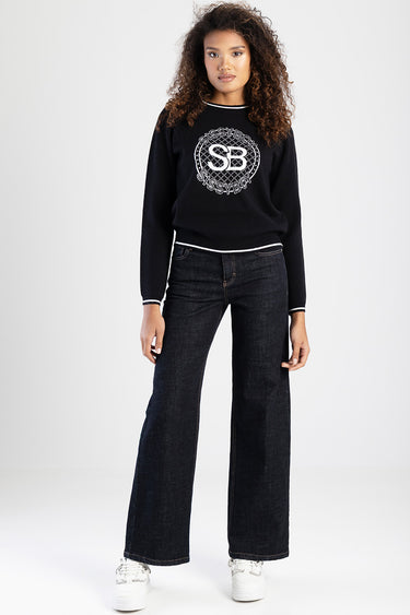 woman wearing a navy knit sweater with a circular logo by blue jean traders, paired with dark wide leg jeans and white