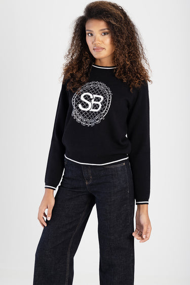 woman wearing a black knit sweater with a white circular logo and white trim at the neckline and cuffs, paired with dark