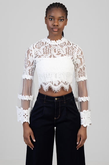 woman wearing a white lace high-neck cropped blouse with sheer long sleeves and scalloped hems, paired with dark trousers.