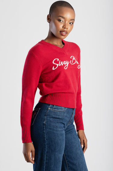 red knit sweater with a v neck and ribbed cuffs and hem, white script logo across the chest, model wearing dark blue jeans