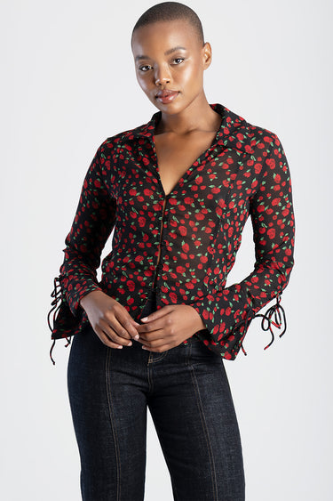 woman wearing a black sheer blouse with red rose print and collared neckline, long sleeves with tie cuffs, paired with dark