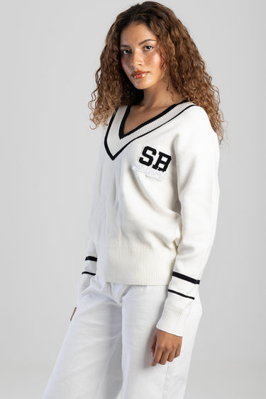 woman wearing blue jean traders white knit sweater with a black v neck trim and sb patch on the chest paired with white pants