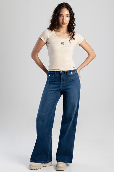 woman in a beige short sleeve top with a small logo standing in wide leg blue denim jeans and beige boots