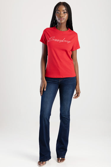 woman wearing a red short sleeve t shirt with a white cursive logo and dark blue jeans, standing against a neutral backdrop