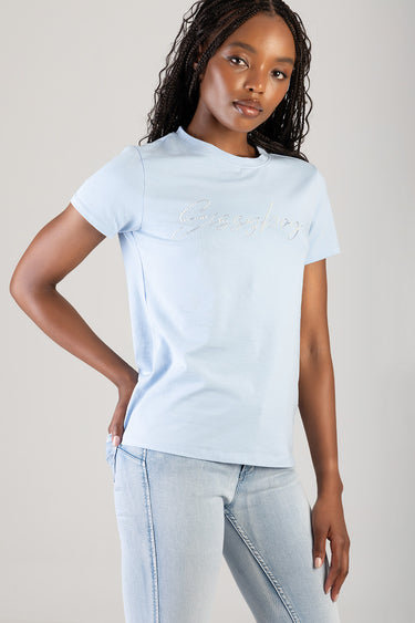 woman wearing light blue short sleeve t shirt with beaded script across chest, standing in light wash jeans.