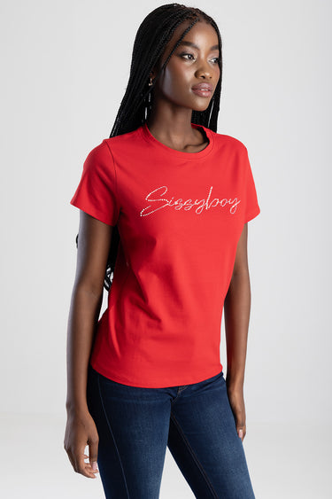 woman wearing a red short sleeve top with rhinestone script across the chest, dark jeans, neutral studio backdrop