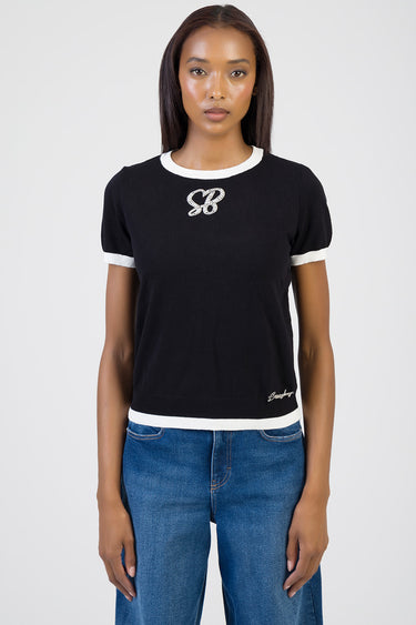 woman wearing black knit top with white trim at neck and cuffs, emblem on chest and small logo at hem, paired with blue jeans