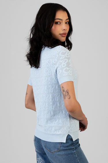 woman in pale blue lace knit top with short sleeves and ribbed hem, standing in profile with blue jeans
