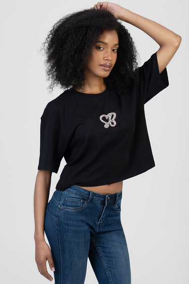 woman wearing a black boxy short sleeve t shirt with a silver rhinestone heart emblem on the chest and blue denim jeans