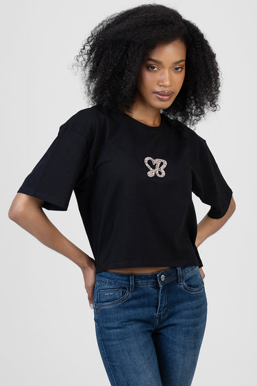 woman wearing a black cropped boxy t-shirt with short sleeves and a rhinestone heart logo on the chest, paired with blue