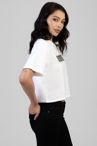 side view of a woman wearing a white boxy short sleeve tee with a rhinestone logo on the chest, paired with black jeans