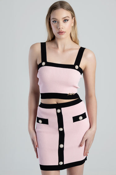 model wearing pink knit crop top with black straps and pearl buttons, paired with a pink skirt with black trim and pearl