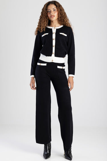 model wearing a black knit cardigan with white trim and gold buttons, paired with matching black trousers with white pocket