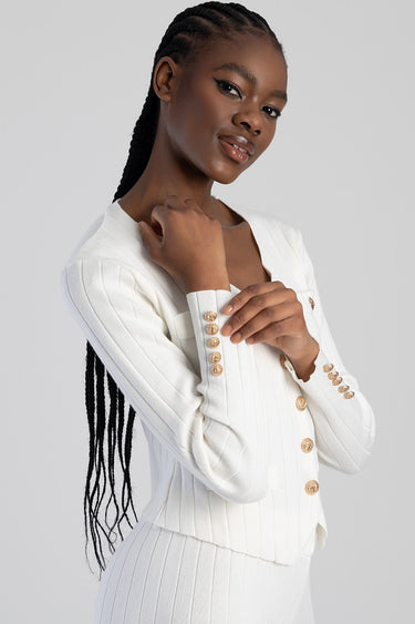 woman wearing a white ribbed knit cardigan with gold buttons, long braids, subtle smile.