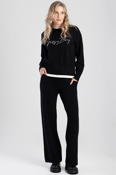 woman wearing a black knit sweater with a white hem and cursive logo, paired with black wide leg pants and chunky black