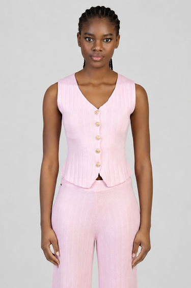 woman in a light pink knit waistcoat with a v neck, gold buttons and ribbed texture, paired with matching trousers.