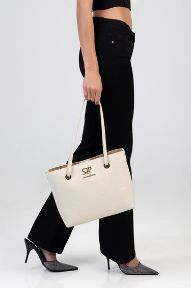 beige tote bag with embossed floral pattern and gold logo, carried by a person in black pants and pointed heels