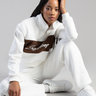 woman seated wearing white track pants and a white zip sweater with a brown panel across the chest; white sneakers