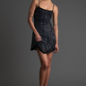 woman in a black sequined mini dress with thin straps and a scattered white dot pattern, standing against a grey backdrop