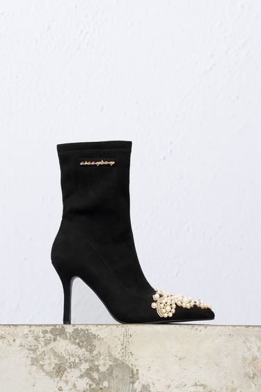 black suede sock boot with a slim heel and pearl embellishment at the toe, gold logo plaque near the top