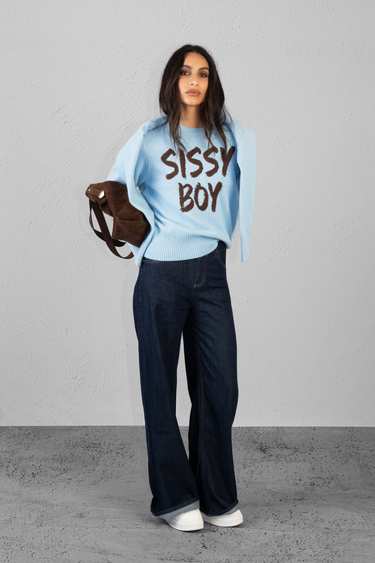 woman wearing a light blue knit sweater with bold dark brown letters, dark wide leg jeans, white sneakers, carrying a brown